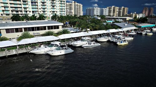 A marina with several boats docked alongside a waterfront restaurant, surrounded by palm trees and city buildings.