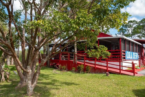 A red building with a porch and signage reading "Cold Beer," surrounded by trees and green grass.
