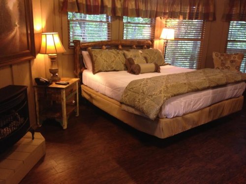 Cozy bedroom with a large bed, decorative pillows, wooden accents, and warm lighting from lamps and windows.