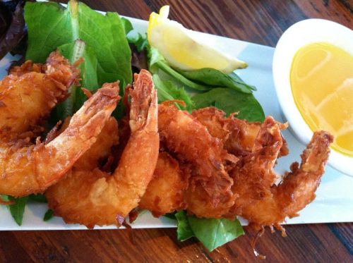 A plate of crispy fried shrimp served with a lemon wedge and a small bowl of dipping sauce on a bed of greens.