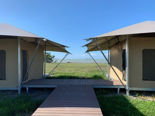 Two glamping tents with a wooden walkway between them, overlooking a grassy area and a calm body of water under a clear blue sky.