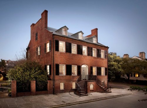 Historic brick house with large windows and a staircase, illuminated at dusk, surrounded by trees and a quiet street.