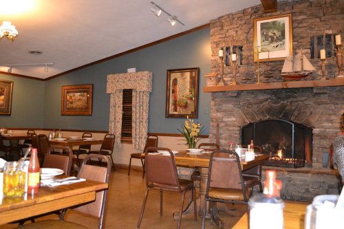 Cozy restaurant interior with wooden tables, chairs, stone fireplace, and framed artwork on the walls.