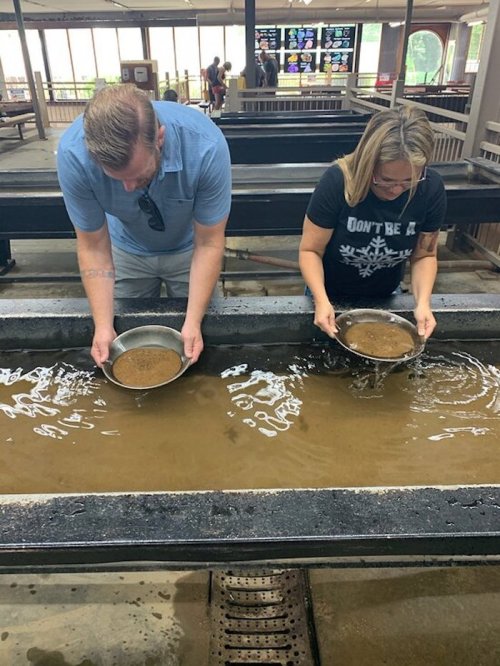 Two people are panning for gold in shallow water, each holding a pan filled with sediment.