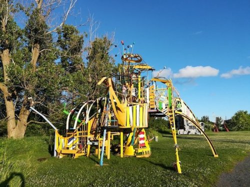 Colorful playground structure with slides and climbing features, surrounded by green grass and trees under a blue sky.
