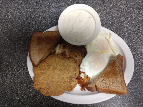 A plate with two fried eggs, hash browns, a bread slice, a chicken patty, and a bowl of gravy.