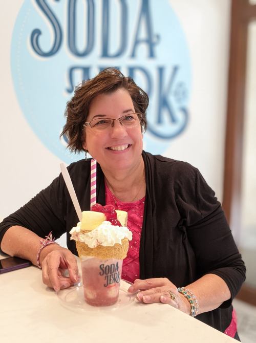 A smiling woman holds a large, colorful soda float topped with whipped cream and a cherry, in front of a "Soda Jerk" sign.