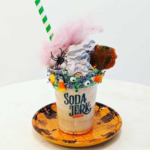 A whimsical milkshake topped with cotton candy, candy corn, and a spider, served in a decorative cup on a Halloween plate.