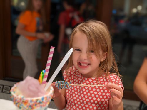 A smiling girl with long hair holds a colorful treat with candy, wearing a polka dot shirt, in a lively setting.