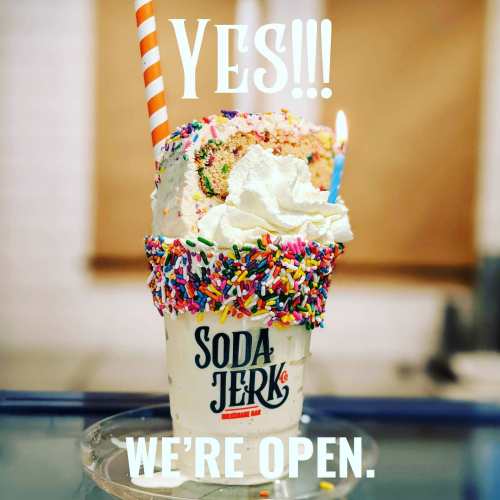 A colorful milkshake topped with whipped cream, sprinkles, and a slice of cake, with a sign saying "YES!!! WE'RE OPEN."