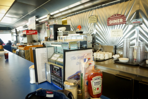 A retro diner counter with a jukebox, condiments, and a menu, featuring a classic chrome design and vintage signage.