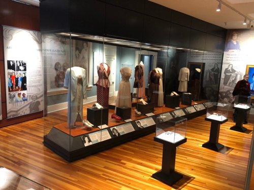 Exhibit showcasing various historical garments displayed in glass cases on a wooden floor.
