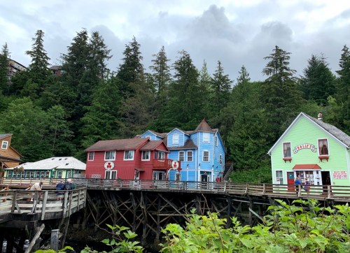 Colorful buildings on stilts by a forested hillside, with a cloudy sky above and a wooden walkway in the foreground.