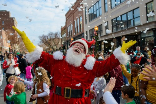 Santa Claus joyfully spreads holiday cheer as snow-like confetti falls around him, surrounded by excited children and families.