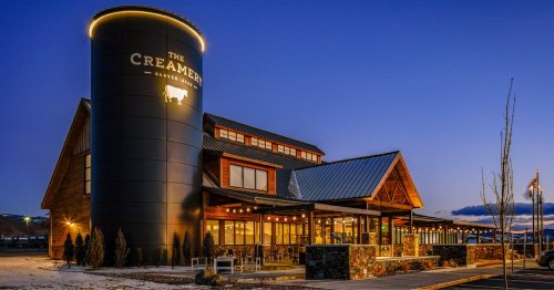 A modern restaurant with a cylindrical tower, wooden exterior, and outdoor seating, illuminated at dusk.