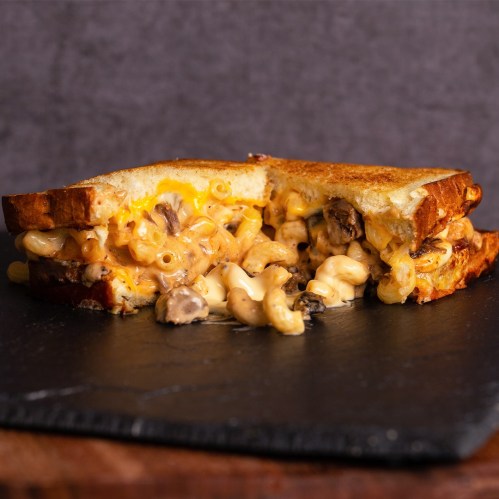 A grilled cheese sandwich filled with creamy macaroni and cheese, featuring mushrooms, on a dark slate surface.