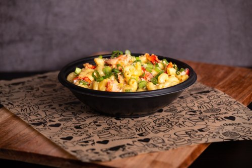 A bowl of creamy macaroni and cheese topped with green onions and diced tomatoes, placed on a patterned paper.