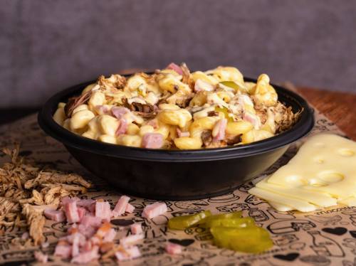 A bowl of creamy macaroni and cheese topped with ham, alongside slices of cheese and pickles on a patterned surface.