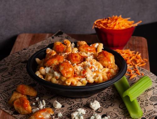 A bowl of macaroni and cheese topped with crispy chicken, buffalo sauce, and garnished with celery and shredded cheese.