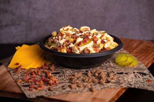 A bowl of loaded nachos topped with cheese, corn, bacon bits, and pickles, served on a decorative paper.
