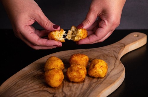 Hands pulling apart a cheesy, golden-brown snack, with more snacks on a wooden serving board.