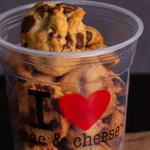 A clear container filled with chocolate chip cookies, with one cookie resting on top, featuring a heart logo.