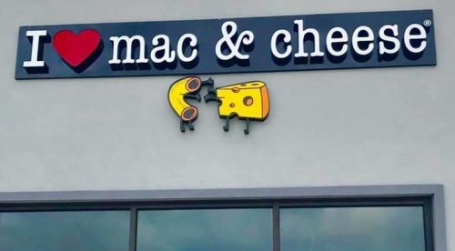 A woman stands in front of a restaurant sign that says "I ❤️ mac & cheese," pointing excitedly.