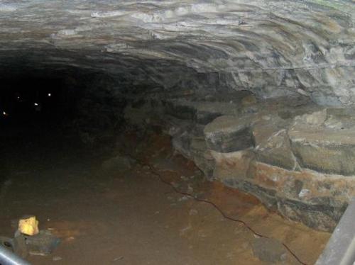 A dimly lit underground tunnel with stone walls and a dirt floor, featuring faint lights in the distance.
