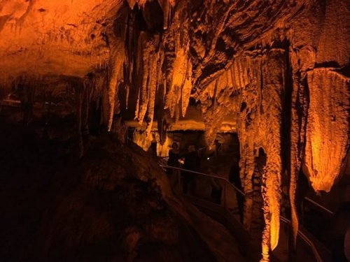 Stalactites and stalagmites illuminated in warm orange light inside a cave, with visitors exploring the area.