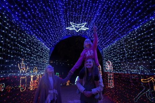 Two girls enjoy a festive light display, one on the other's shoulders, reaching for a star-shaped light above.