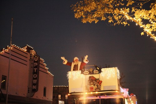 Santa waves from the top of a decorated fire truck during a nighttime holiday parade, with festive lights in the background.