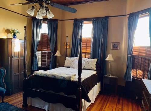 Cozy bedroom with a four-poster bed, blue curtains, wooden floors, and warm lighting from windows.