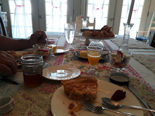 A table set with various dishes, including pie, fruit, and drinks, with hands reaching for food.