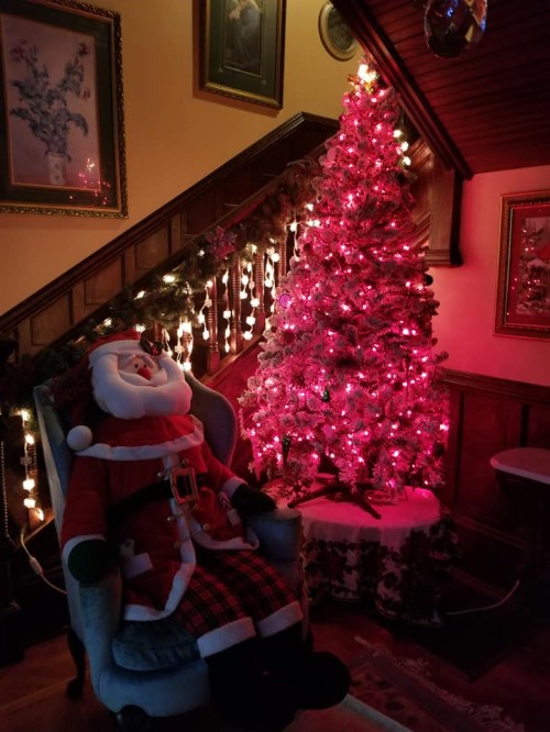 A cozy holiday scene featuring a Santa figure sitting in a chair next to a pink-lit Christmas tree and festive decorations.