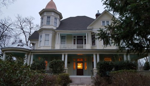 A large, historic Victorian house with a turret and porch, surrounded by trees and shrubs, at dusk.