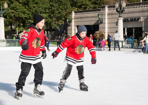 Two boys in red hockey jerseys skate on an ice rink, surrounded by trees and people enjoying the day.
