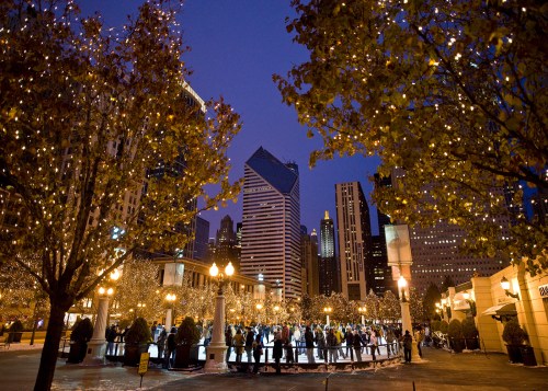 A bustling outdoor ice skating rink surrounded by twinkling lights and city skyscrapers at dusk.