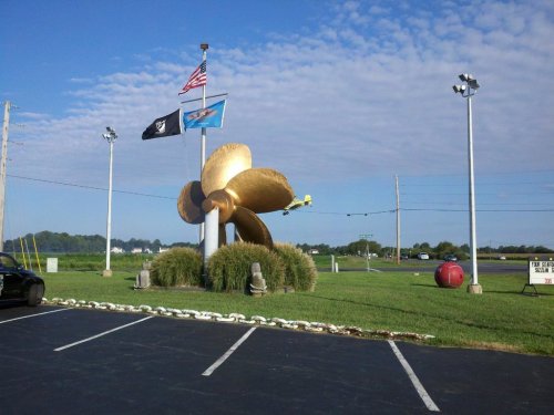 A large golden propeller sculpture stands in a grassy area, with flags and a clear blue sky in the background.