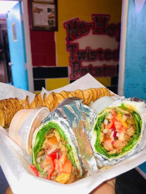 A hand holding a tray with two wrapped sandwiches and a side of crispy potato chips. Colorful wall in the background.