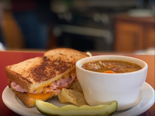 A toasted sandwich with ham and cheese, served with a bowl of soup and a pickle on the side.