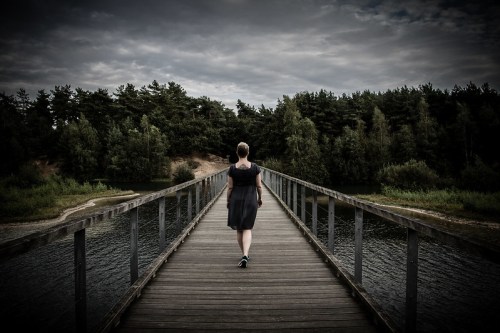 A person walks alone on a wooden bridge over a calm body of water, surrounded by trees under a cloudy sky.