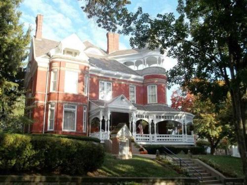 A large red brick Victorian house with a white porch, surrounded by trees and greenery under a blue sky.