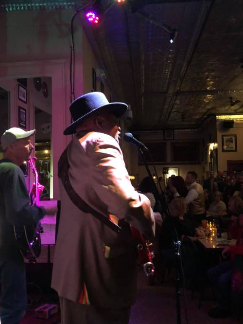 A musician in a hat performs on stage, with a guitarist and a lively audience in a dimly lit venue.