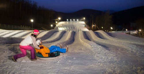 A person in a bright orange jacket is being pushed on a snow tube down a snowy slope at night, with lights in the background.