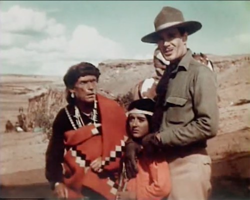 A man in a hat stands with a woman and an older man, all dressed in traditional attire, against a desert landscape.