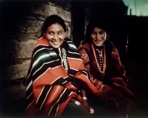 Two smiling women wrapped in colorful blankets, sitting against a stone wall in a dimly lit setting.
