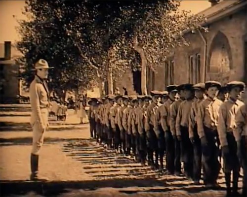 A line of uniformed boys standing in formation outdoors, with an officer supervising in front of a historic building.