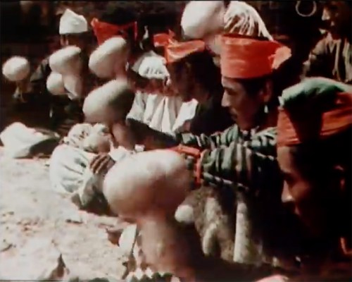 A group of people in traditional attire rhythmically shaking large, round objects during a cultural event.