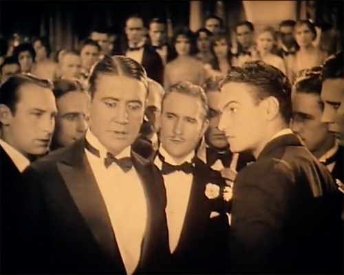 A group of men in tuxedos, gathered closely, engaged in a serious conversation at a formal event.