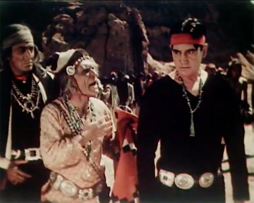 Three men in traditional attire stand in a desert landscape, engaged in a serious conversation.